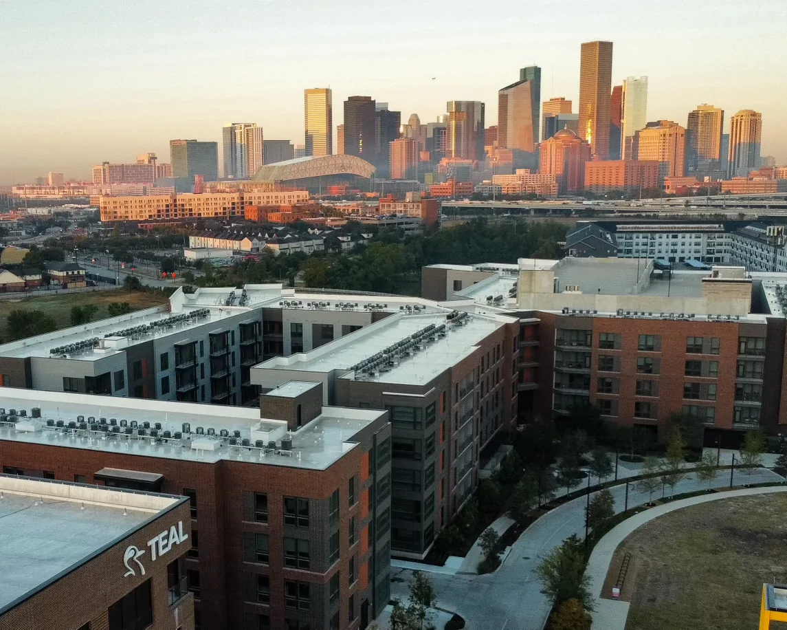 TEAL-branded building and nearby multifamily buildings with the Houston skyline at sunset